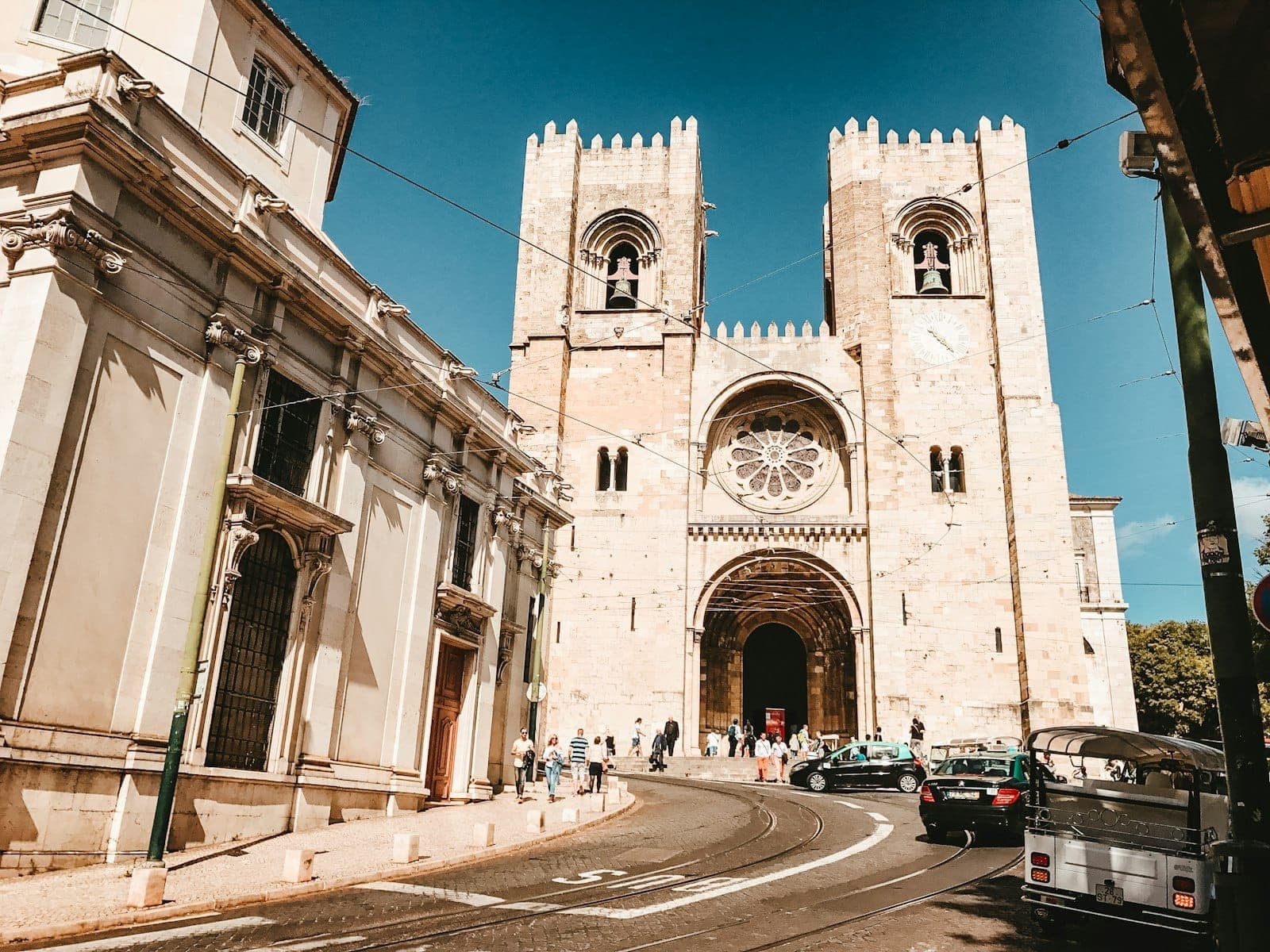 Nhà thờ Sé ở Lisbon với hai tháp chuông và cửa sổ hoa hồng, nhìn thấy vào một ngày nắng đẹp với người và taxi ở phía trước.