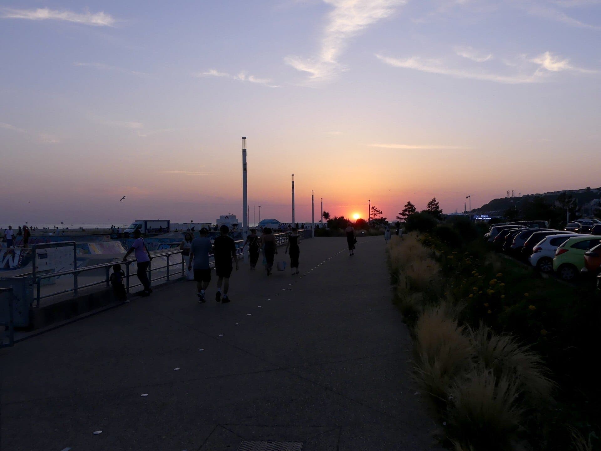 Mọi người dạo bộ dọc theo Promenade Maritime của Le Havre lúc hoàng hôn, với những chiếc xe đậu bên đường, lan can ven biển và ánh sáng buổi tối dịu dàng nhuộm màu bầu trời.