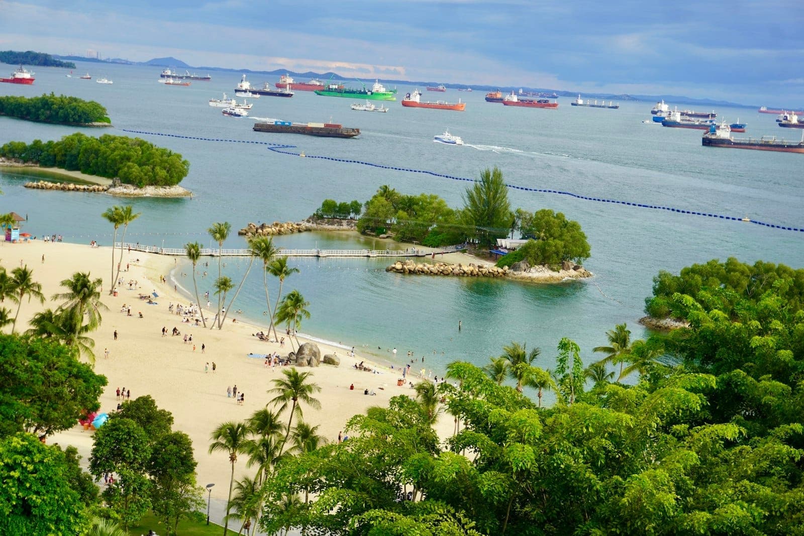 A panoramic view of Sentosa Island’s beach with golden sand, palm trees, small offshore islets, and ships anchored in the background under a blue sky.