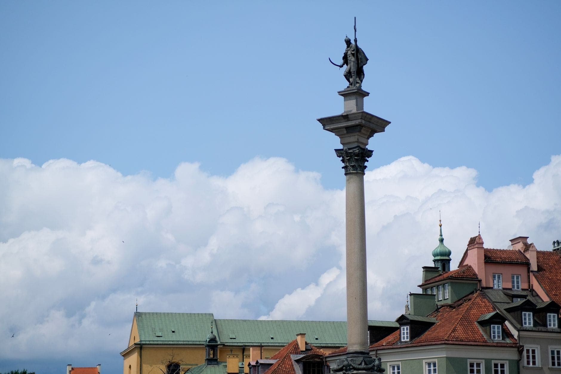 Die Sigismundsäule erhebt sich über den bunten Gebäuden auf dem Schlossplatz in Warschau vor einem strahlend blauen Himmel mit weißen Wolken.
