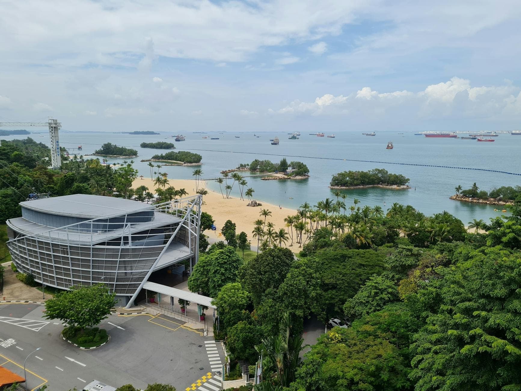 Aerial view of Sentosa Island with sandy beaches, palm trees, a modern building, and ships anchored off the coast.