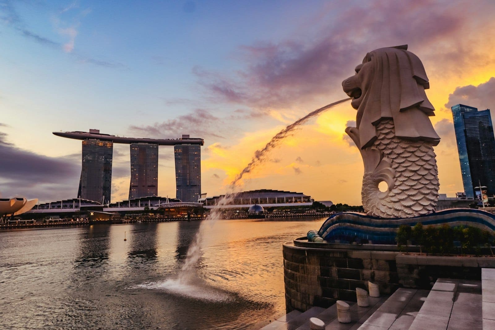 Merlion statue in front of Marina Bay Sands at sunset, with dramatic sky and city skyline, inviting viewers to explore Singapore and nearby escapes.