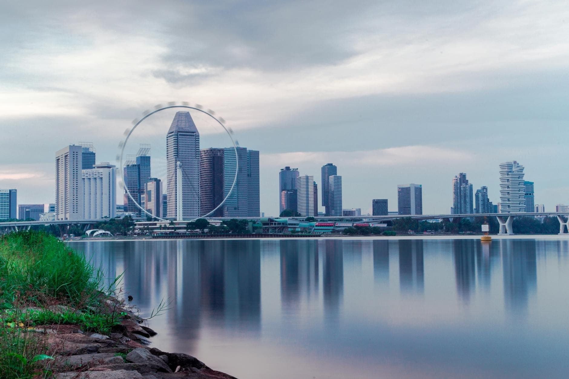 View of the Singapore Flyer observation wheel towering above the Marina Bay city skyline, reflected peacefully in the calm waterfront at dusk.