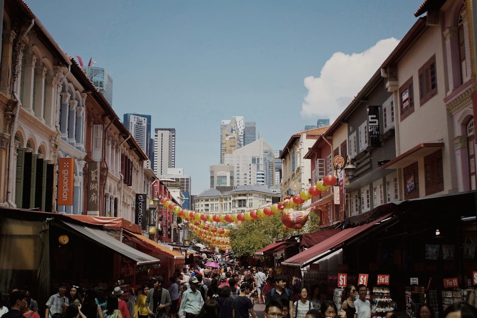 Busy Chinatown street in Singapore with colorful lanterns and heritage shophouses, filled with people and city skyscrapers visible in the background.