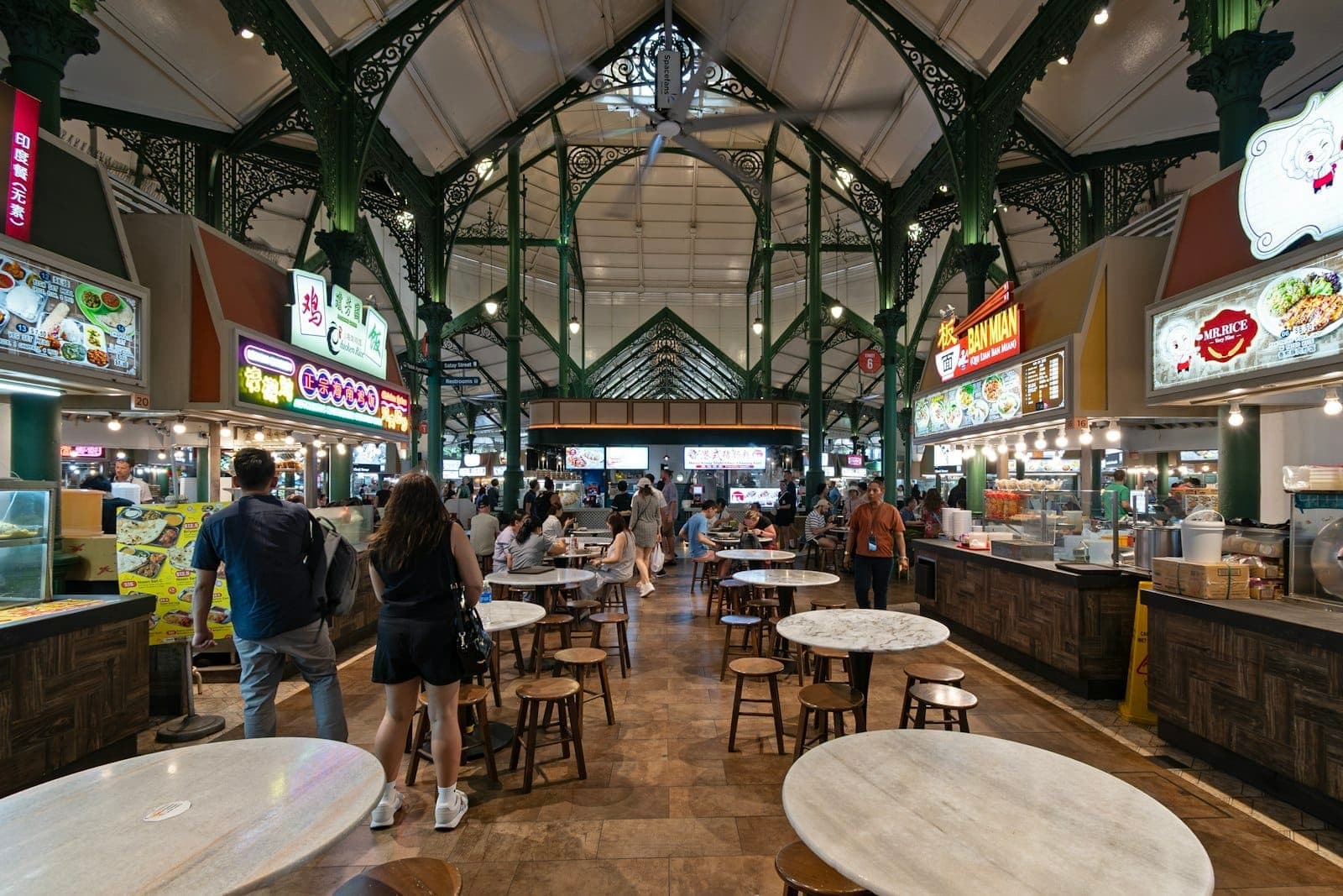 Wide angle view inside a bustling Singapore hawker centre with marble tables, food stalls, and people exploring different local dishes.