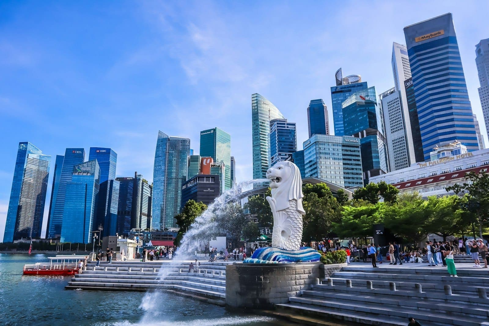 Singapore’s iconic Merlion statue with water spouting, dramatic skyline of modern skyscrapers, and vibrant crowds under a sunny blue sky at Marina Bay.