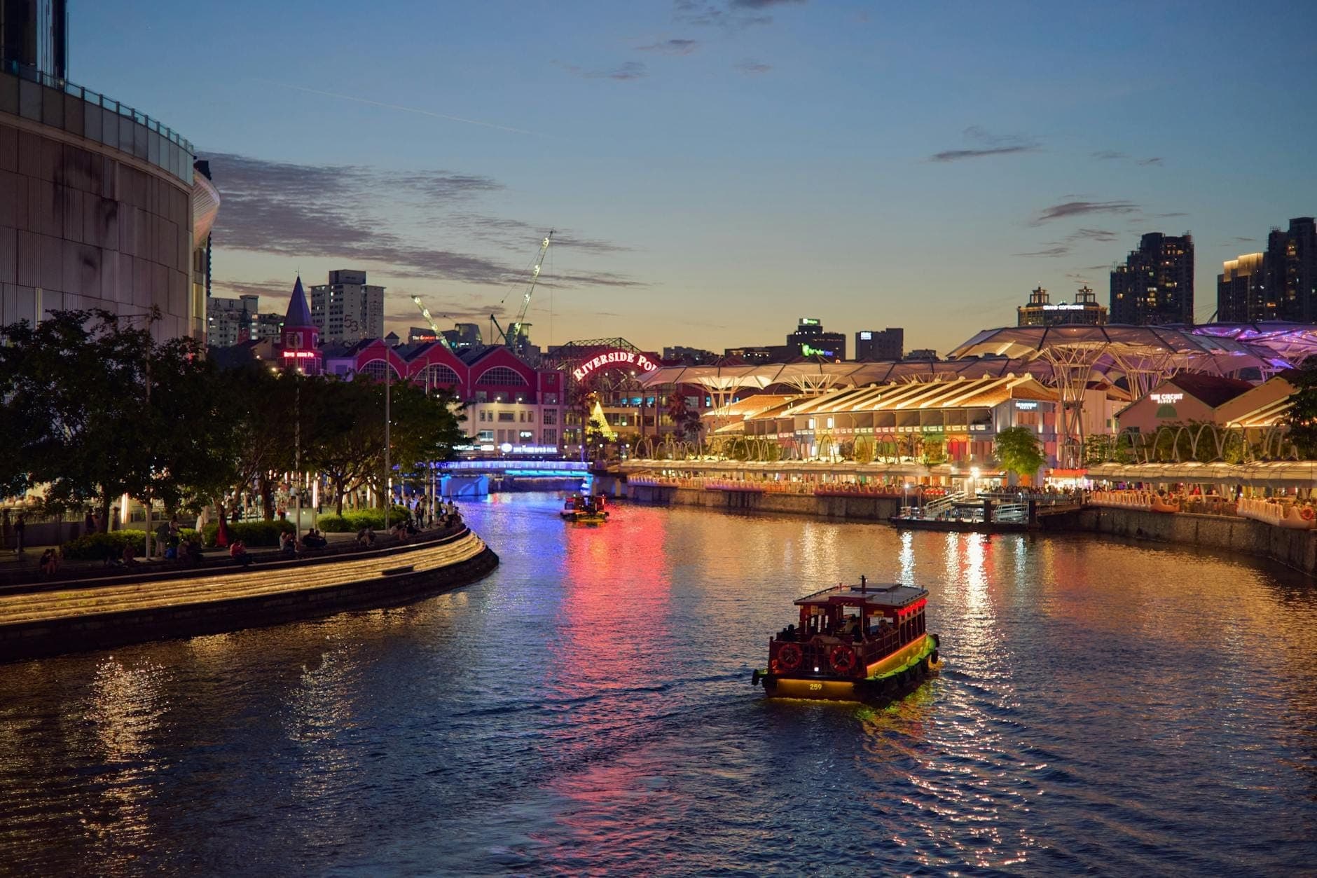 Traditional bumboat cruising along Singapore River at dusk, with Clarke Quay’s vibrant lights and modern city skyline creating a lively waterfront atmosphere.