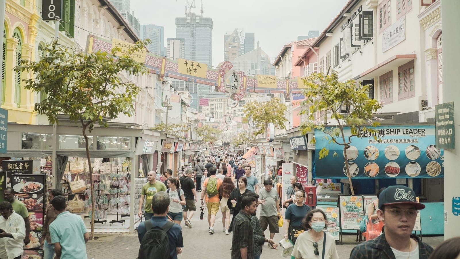 Busy Singapore street market with colorful shopfronts, crowds of shoppers, and skyscrapers in the background on a bright day.