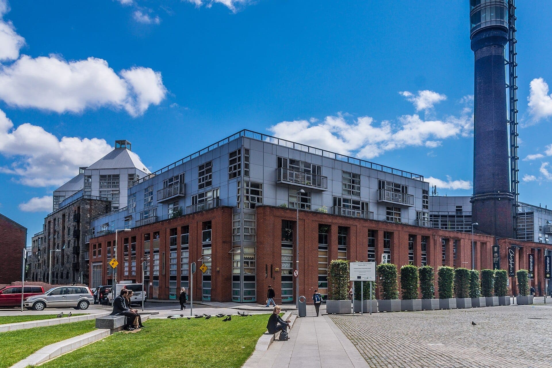 Moderne Backstein- und Metallgebäude säumen den Smithfield Square in Dublin, während Menschen unter einem strahlend blauen Himmel auf begrünten Bänken sitzen.