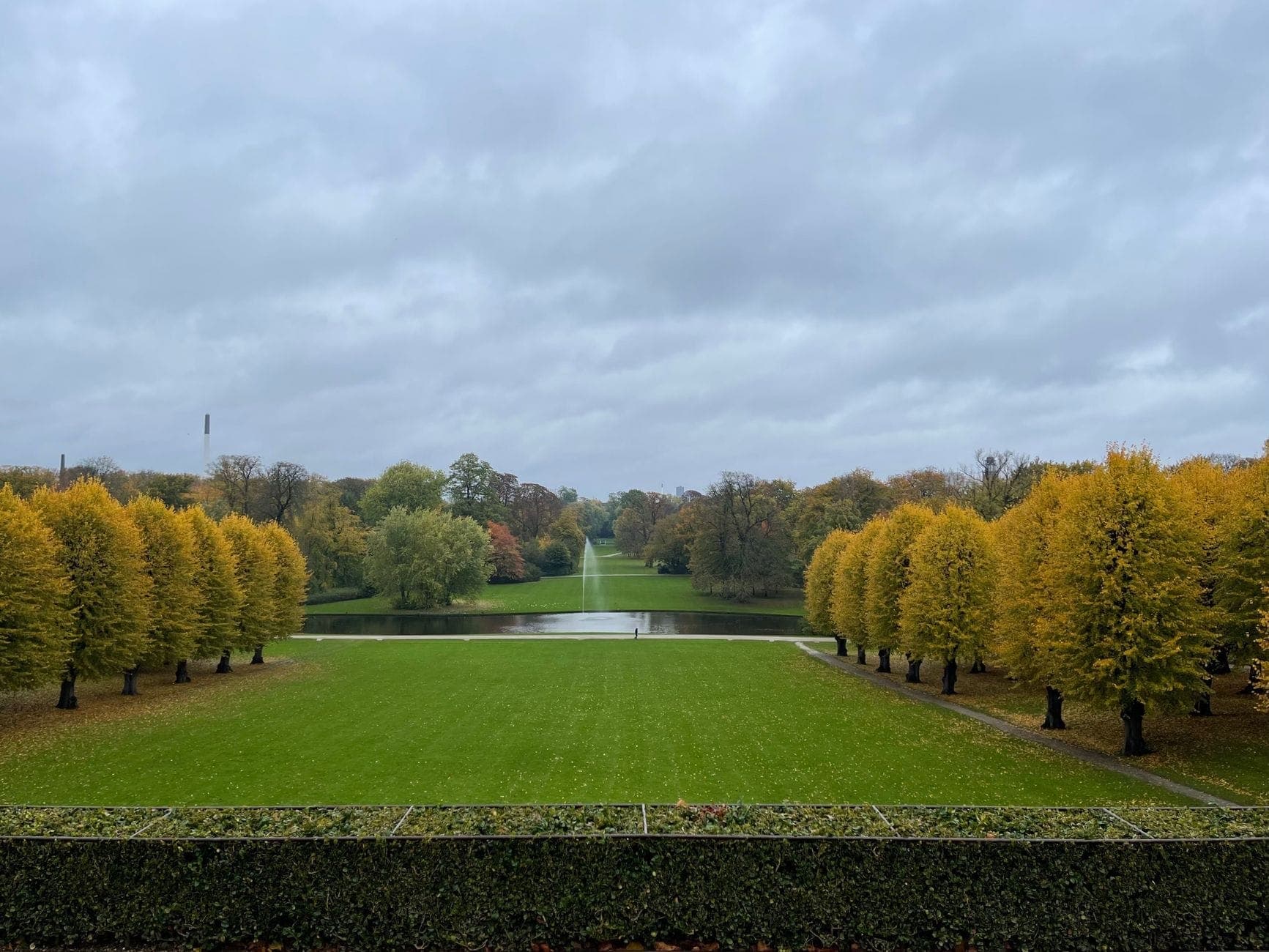 Eine weitläufige Rasenfläche, flankiert von Reihen herbstlich gelber Bäume, mit Waldstücken und einem kleinen Teich unter bewölktem Himmel – eine friedliche Parkszene.