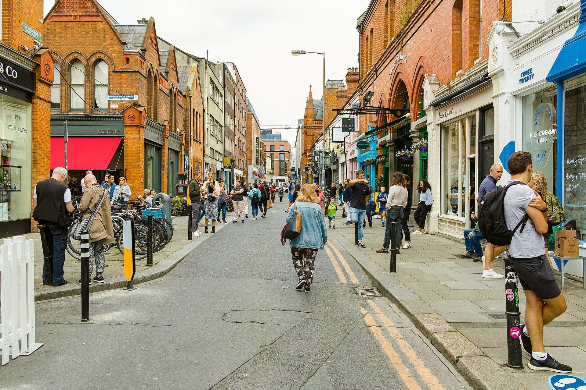 Fußgänger schlendern entlang der South William Street in Dublin, gesäumt von roten Backsteingebäuden im georgianischen Stil, unabhängigen Geschäften und lebhaften Schaufenstern an einem bewölkten Tag.
