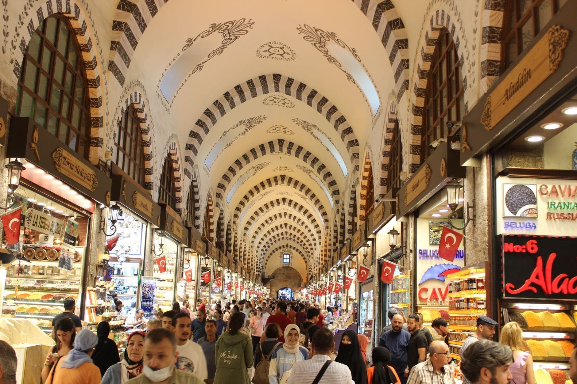 Belebtes Inneres des Istanbuler Gewürzbasars mit gewölbter, verzierter Decke, einkaufenden Menschen und bunten Läden voller Gewürze, Süßigkeiten und türkischer Köstlichkeiten auf beiden Seiten.