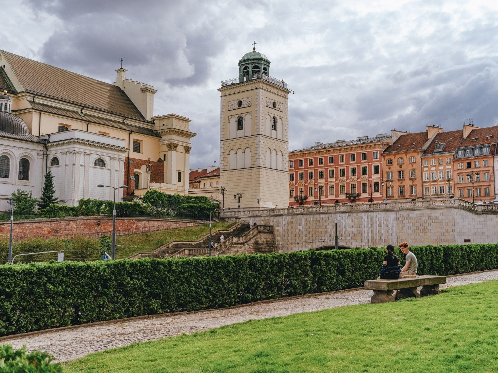 Die St.-Anna-Kirche und ihr markanter Aussichtsturm in Warschau, im Vordergrund Menschen auf einer Bank, sattgrünes Gras und Altstadtgebäude im Hintergrund.
