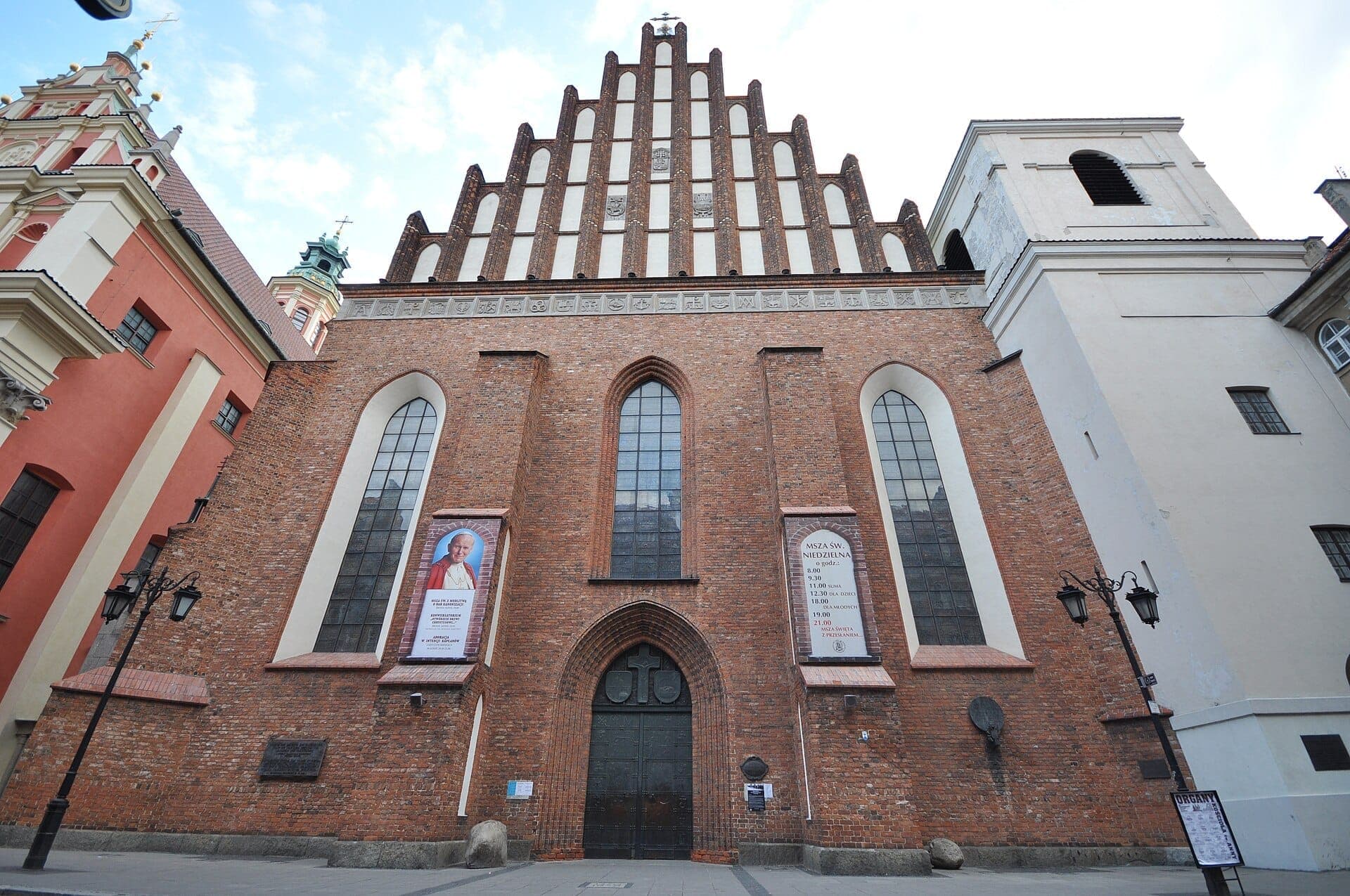 Frontansicht der Erzbischofskathedrale St. Johannes in Warschau mit ihrer hohen Backsteinfassade, gotischen Fenstern und Bannern auf beiden Seiten des Eingangs.