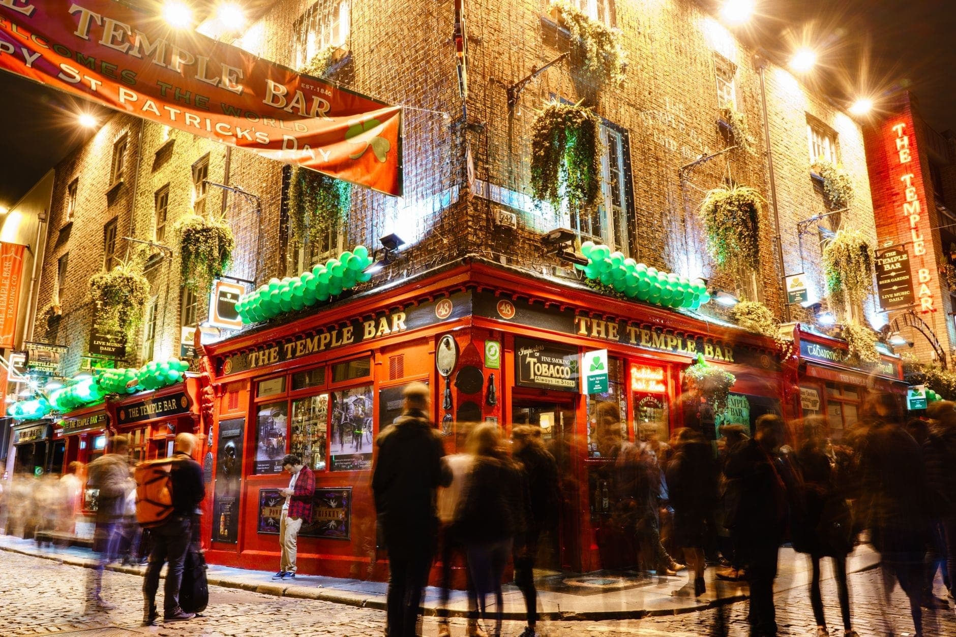 Nachtansicht des Temple Bar in Dublin mit St. Patrick's Day-Bannern, grünen Dekorationen und Menschenmengen vor dem Eingang.