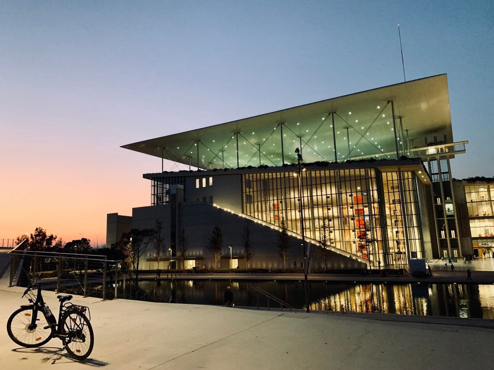 Evening view of Stavros Niarchos Foundation Cultural Center with illuminated glass façade, distinctive angled roof, and reflecting pool, under a colorful sunset sky in Athens.