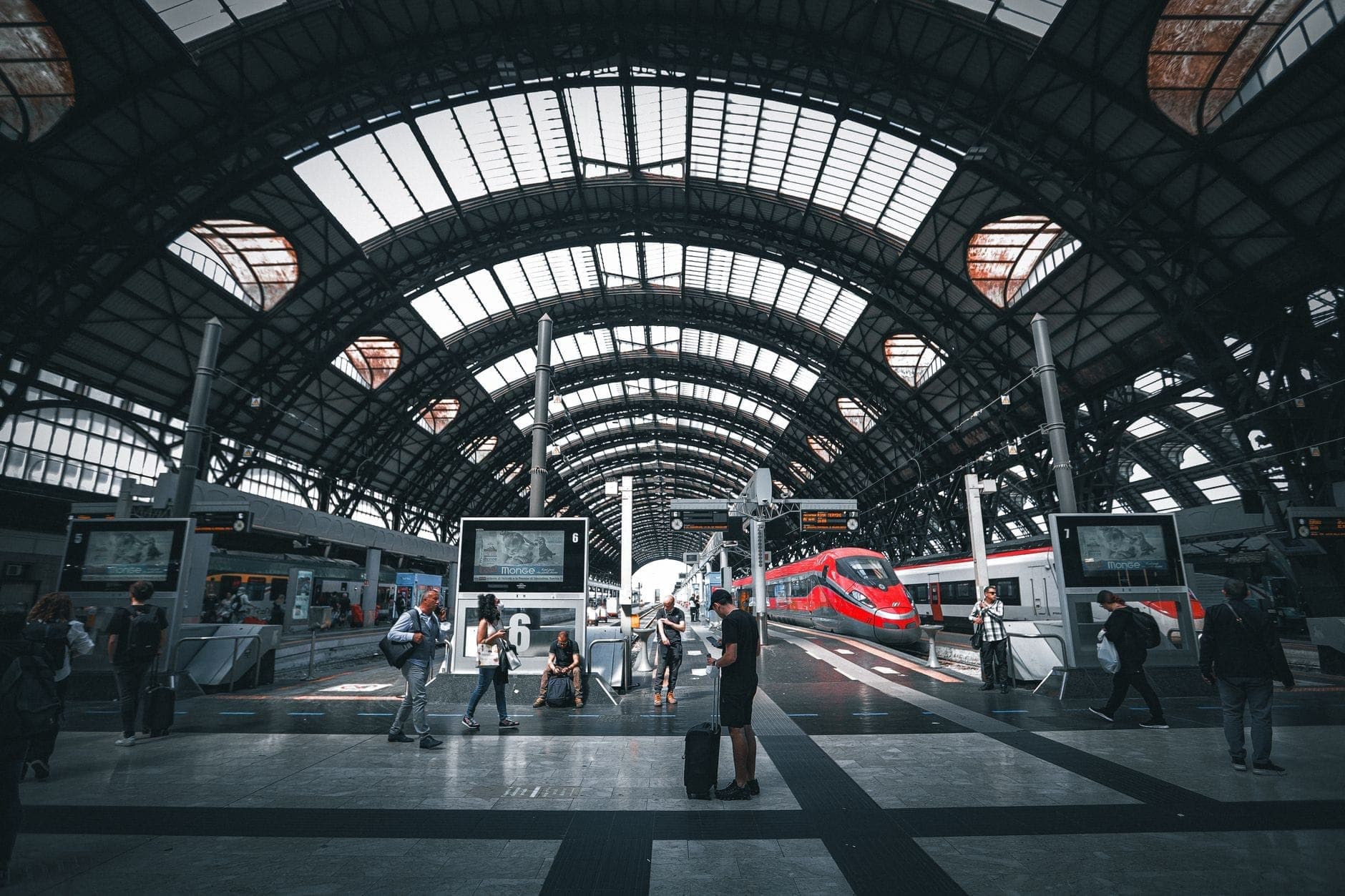 Travelers walk through Milano Centrale Station's expansive glass-and-steel vaulted platform, with a red high-speed train waiting under the arched roof.