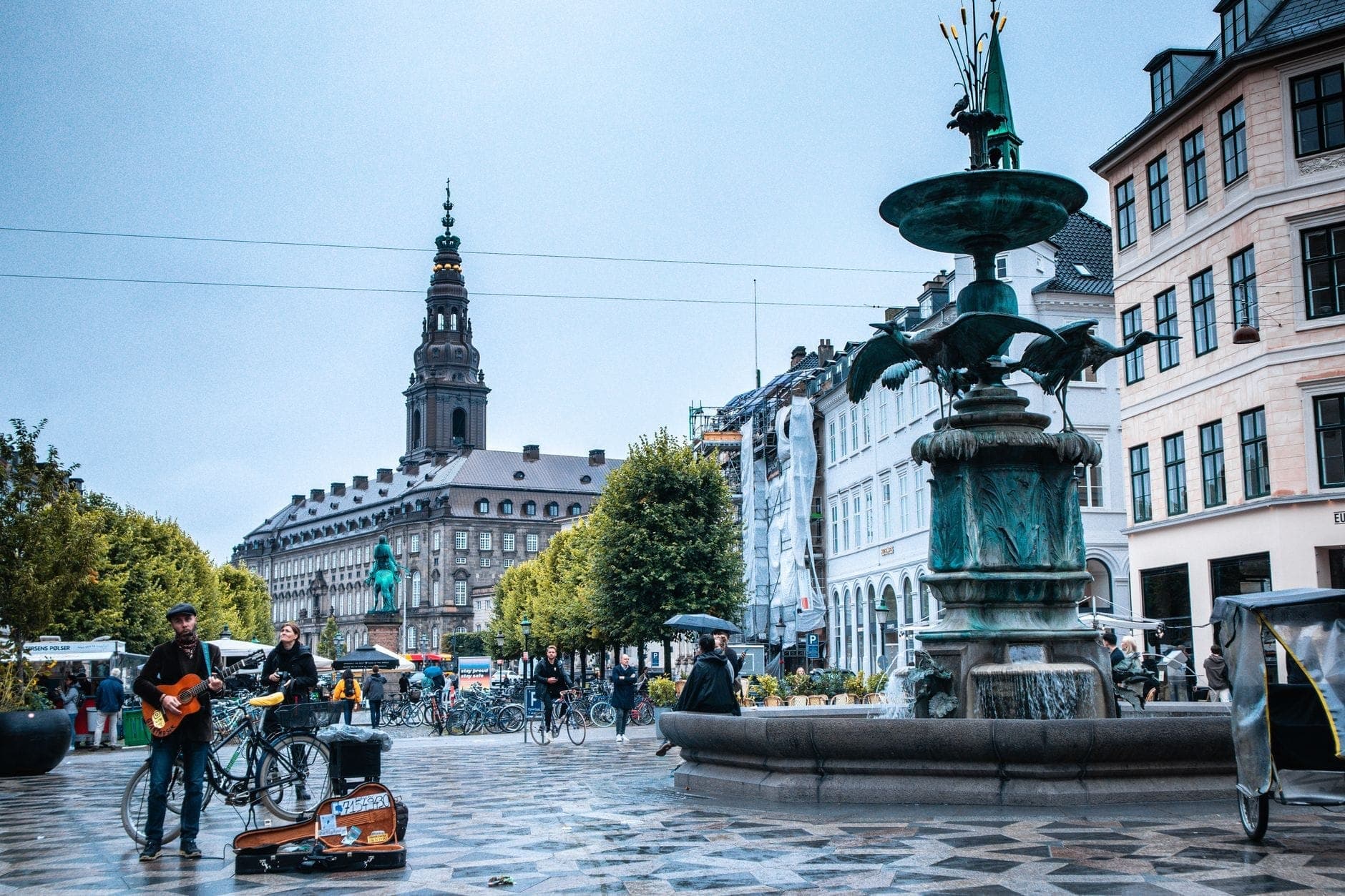 Lebhaftes Treiben auf dem Strøget in Kopenhagen: historische Gebäude, ein großer Brunnen, ein Straßenmusiker mit Gitarre, Fußgänger und Radfahrer.