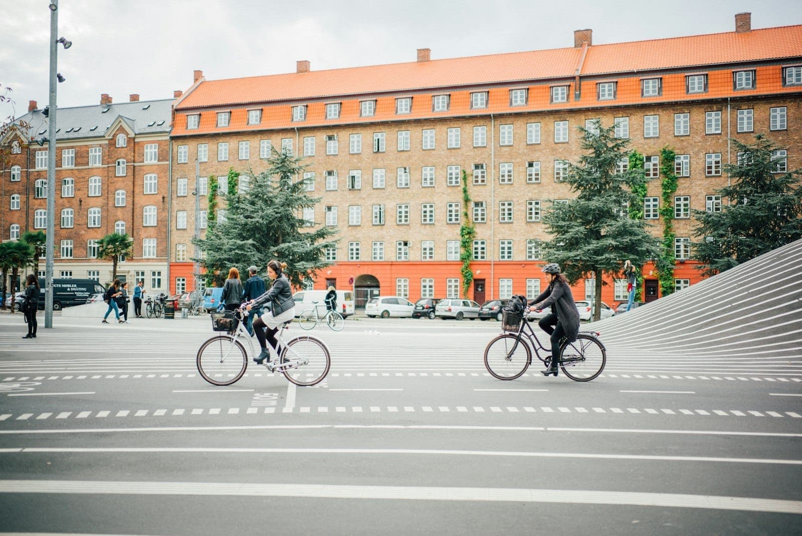 Radfahrer und Fußgänger bewegen sich durch den Superkilen-Park in Kopenhagen, mit gestreiftem Pflaster, immergrünen Bäumen und Backsteinwohnhäusern im Hintergrund.