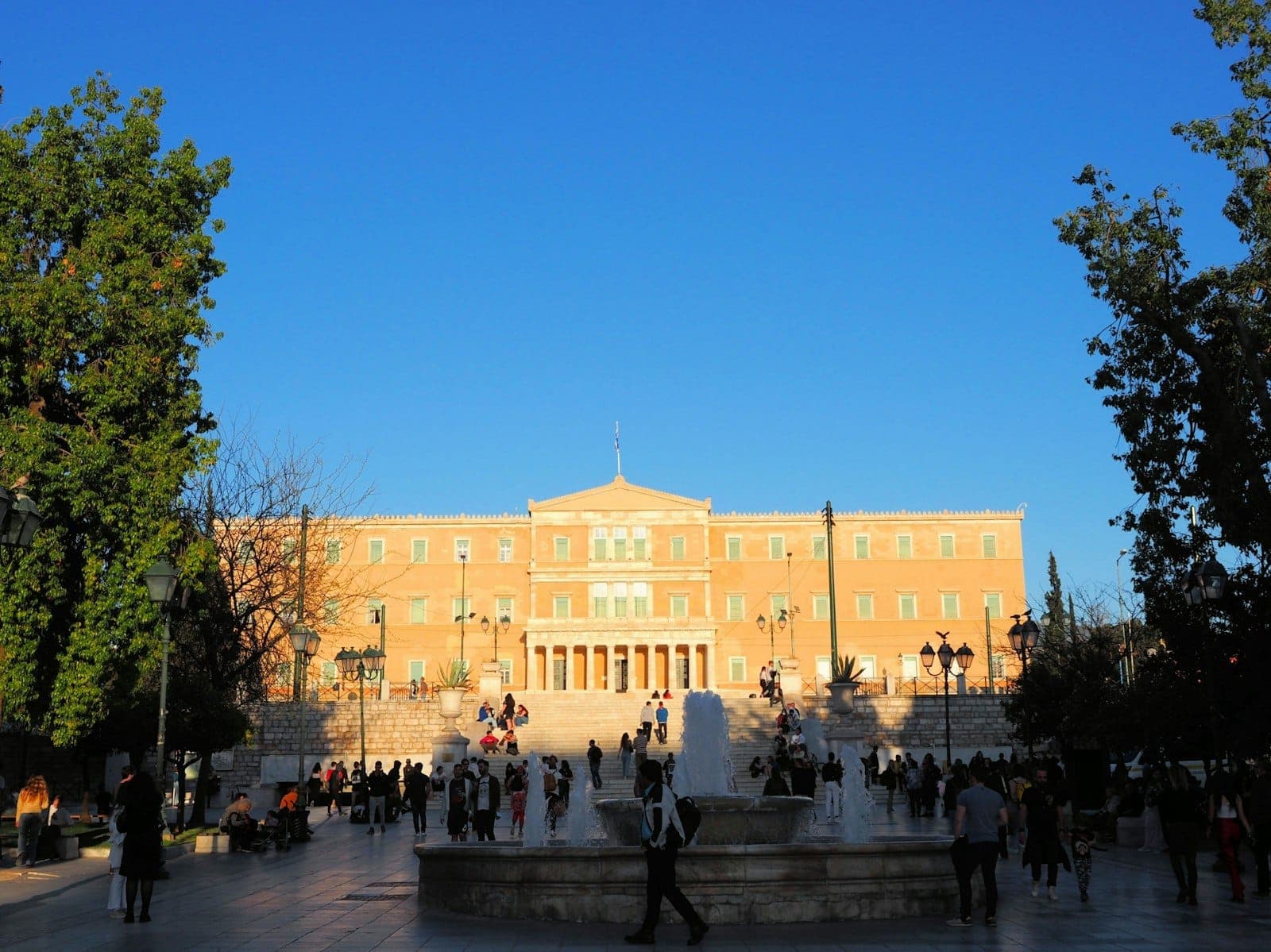 A vibrant scene at Syntagma Square with people gathering around the central fountain, framed by trees, facing the Hellenic Parliament on a clear day.