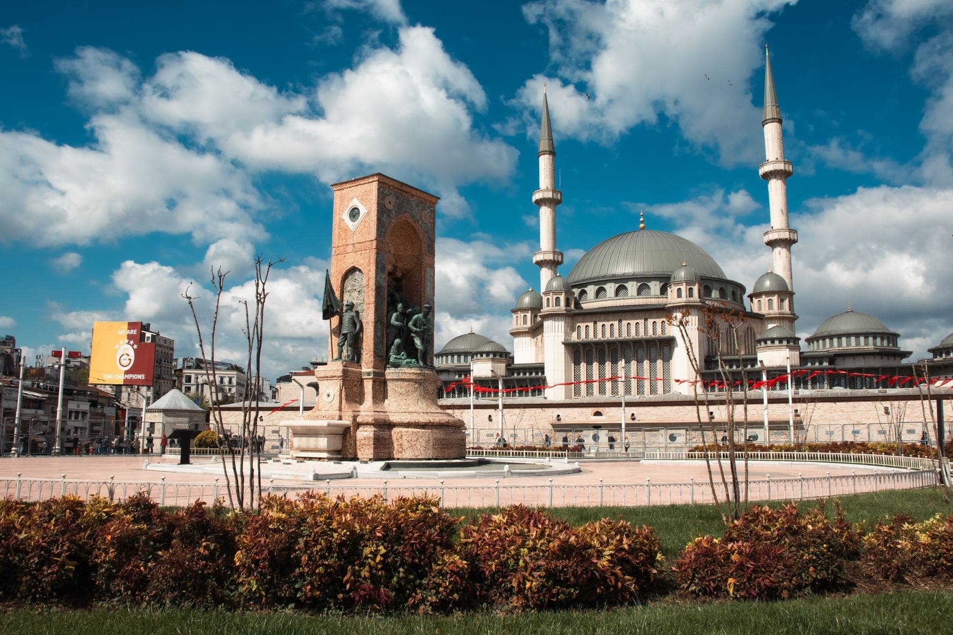 Weiter Blick über den Taksim-Platz mit dem Republikdenkmal in der Mitte und der Taksim-Moschee im Hintergrund unter einem strahlend blauen Himmel mit Wolken.