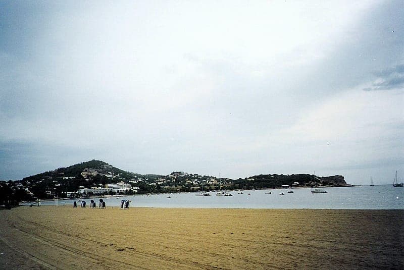 Wide view of Talamanca Beach in Ibiza with sandy shore, calm water, distant buildings, and boats anchored near the coast.
