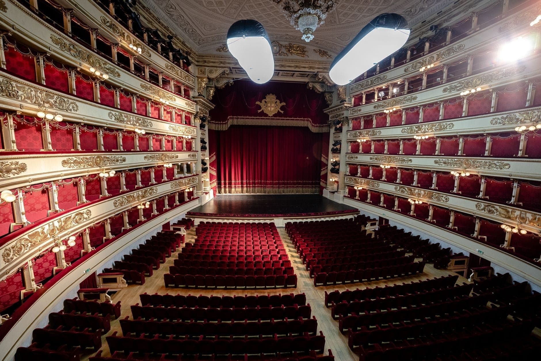 Wide interior view of Teatro alla Scala showing red velvet seats, ornate balconies, and the grand stage with a closed red curtain under elegant lighting.