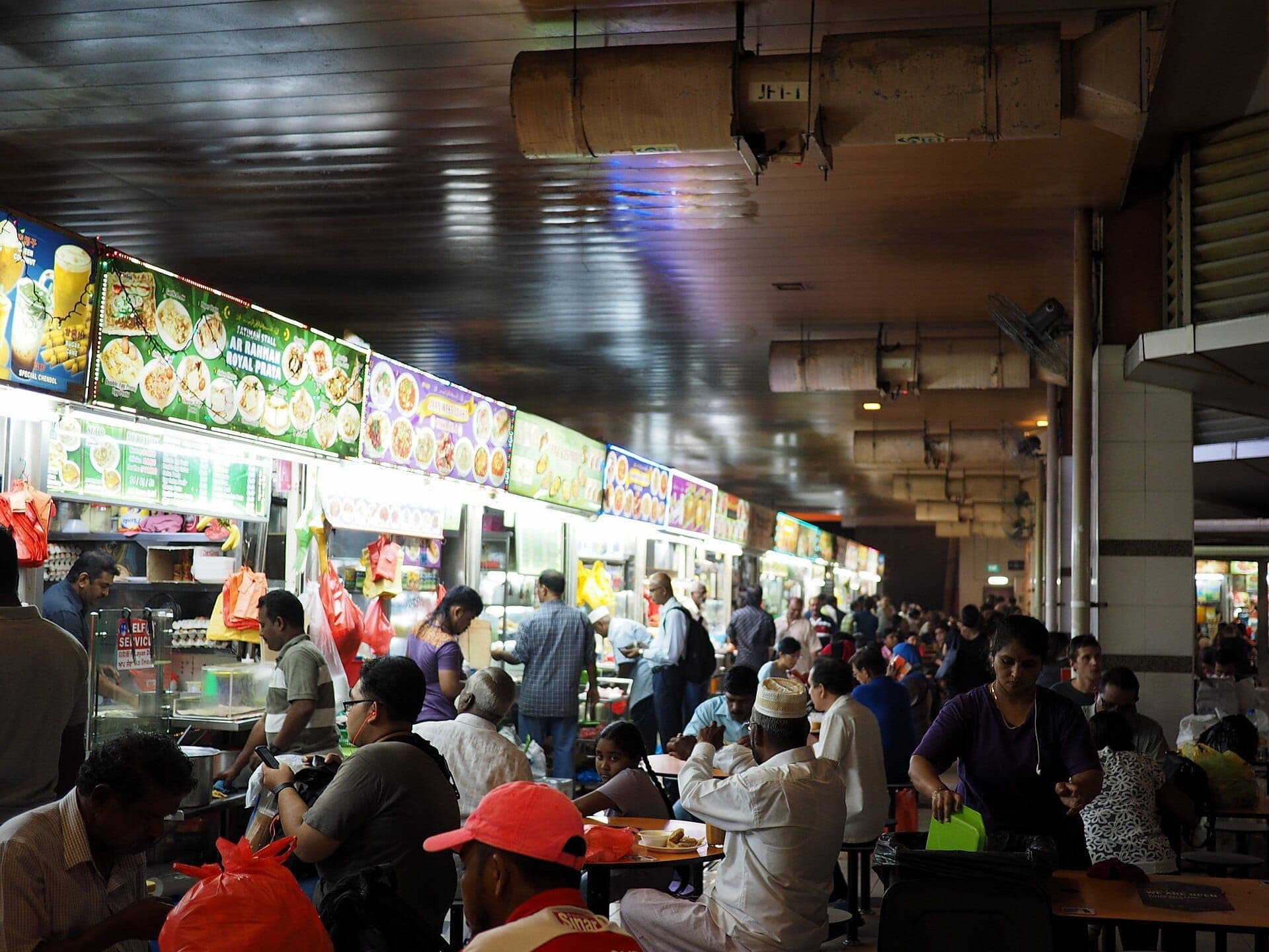 Crowds of people dine and order food at brightly lit hawker stalls inside Singapore’s Tekka Centre, with colorful menus overhead.