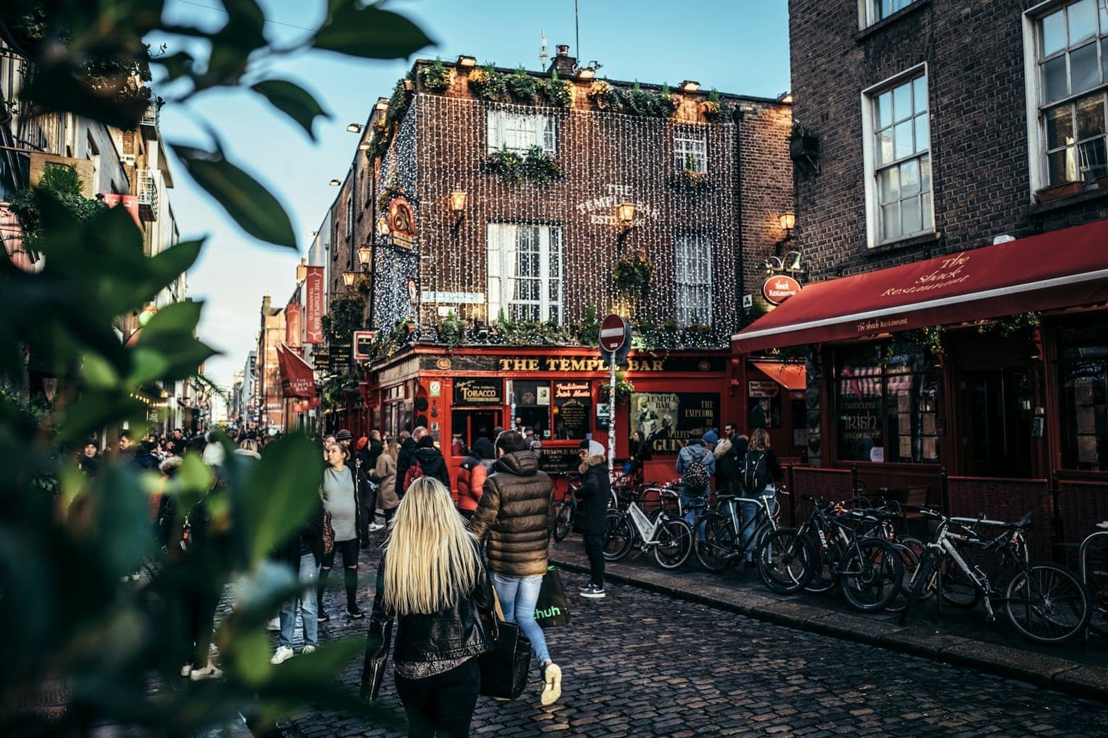 Belebte Kopfsteinpflasterstraße in Temple Bar, Dublin, mit Menschen, die auf den berühmten roten Pub zulaufen, aufgereihten Fahrrädern und festlichen Lichterketten über den Köpfen.