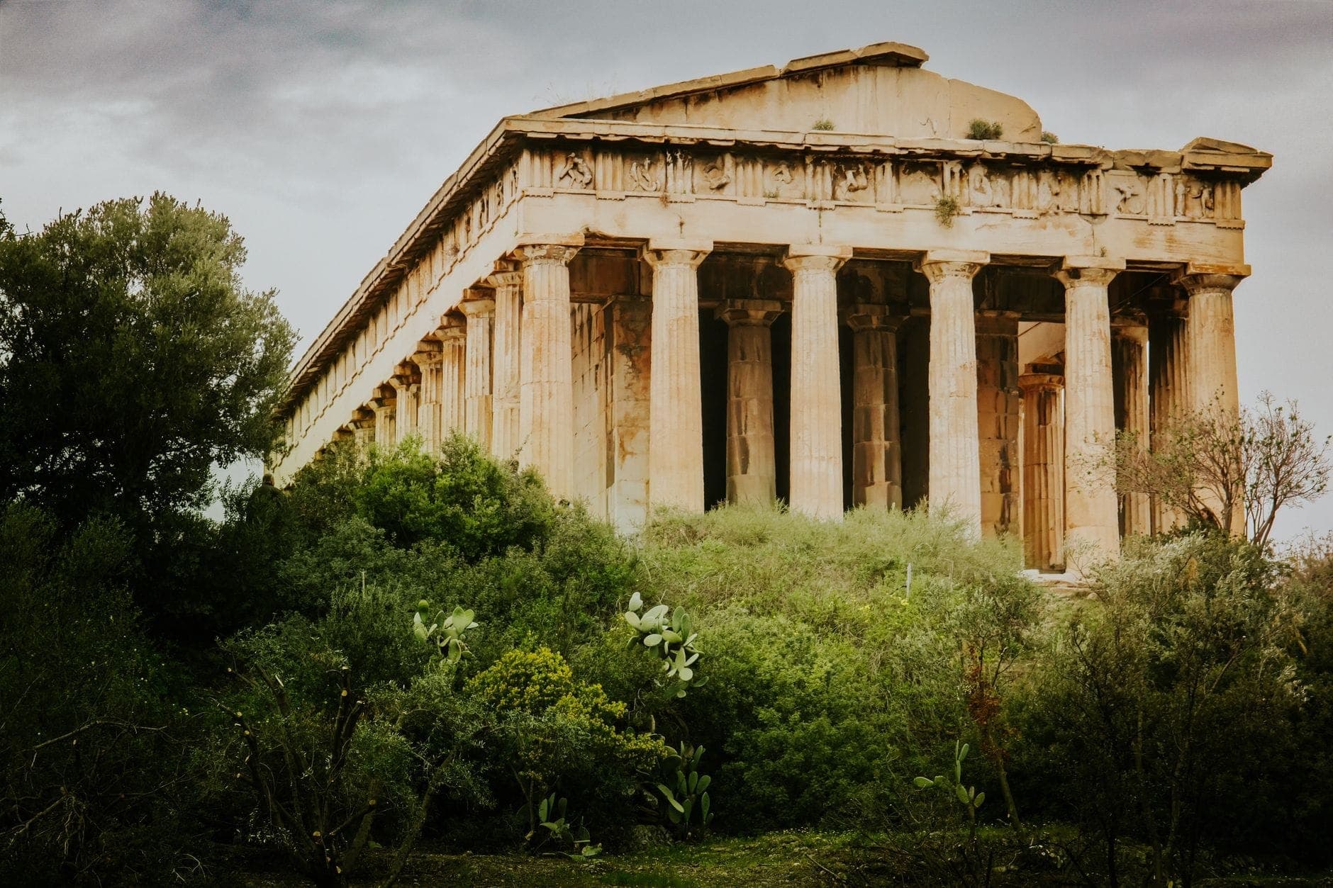 Side view of the well-preserved Temple of Hephaestus in Athens, framed by lush green trees and dramatic cloudy skies, showcasing its iconic Doric columns.