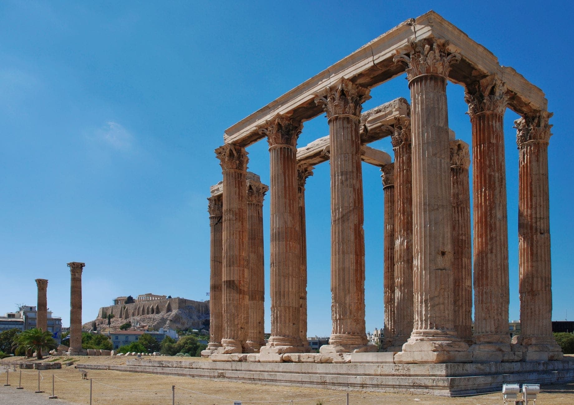 Impressive Corinthian columns of the Temple of Olympian Zeus rise under a blue sky in Athens, with the Acropolis visible in the background.