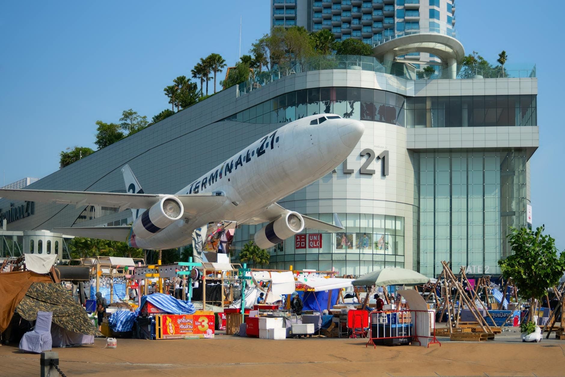 Daytime view of Terminal 21 Pattaya mall with its iconic airplane installation at the entrance and bustling outdoor market stalls in front.