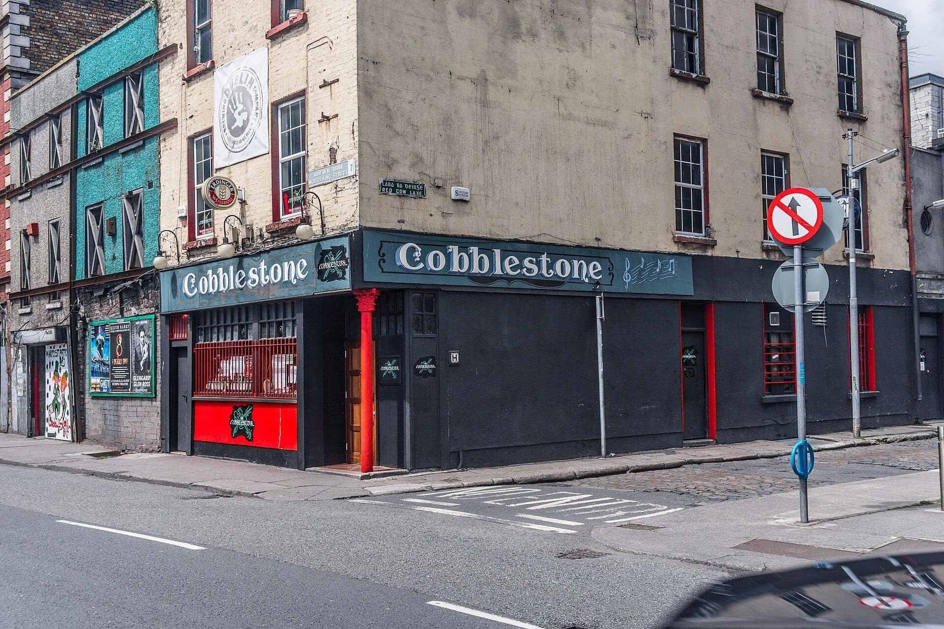 Straßenansicht des Pubs The Cobblestone in Dublin, mit schwarzer Fassade, roten Akzenten und traditionellen Schildern an einem bewölkten Tag.