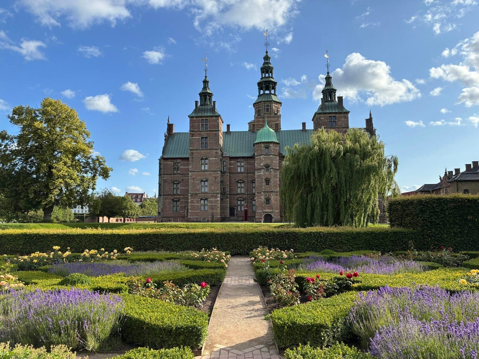Weitläufiger Blick auf den Kongens Have mit farbenfrohen Blumenbeeten und gepflegten Hecken, die zum Schloss Rosenborg unter strahlend blauem Himmel führen.
