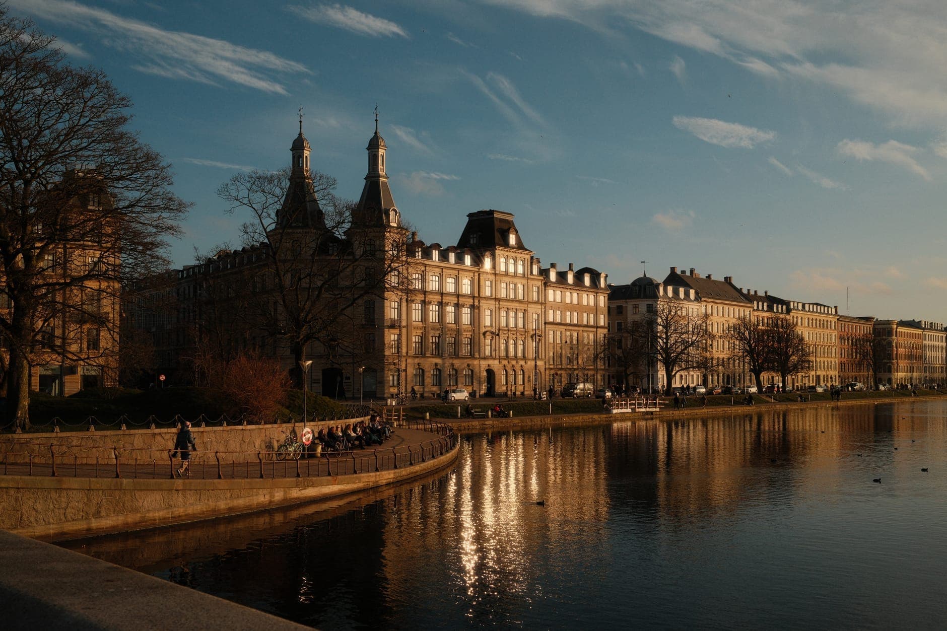 Blick am späten Nachmittag auf Kopenhagens Seeuferpromenade: Menschen spazieren am Wasser entlang, während sich elegante historische Gebäude im ruhigen Wasser unter einem goldenen Himmel spiegeln.