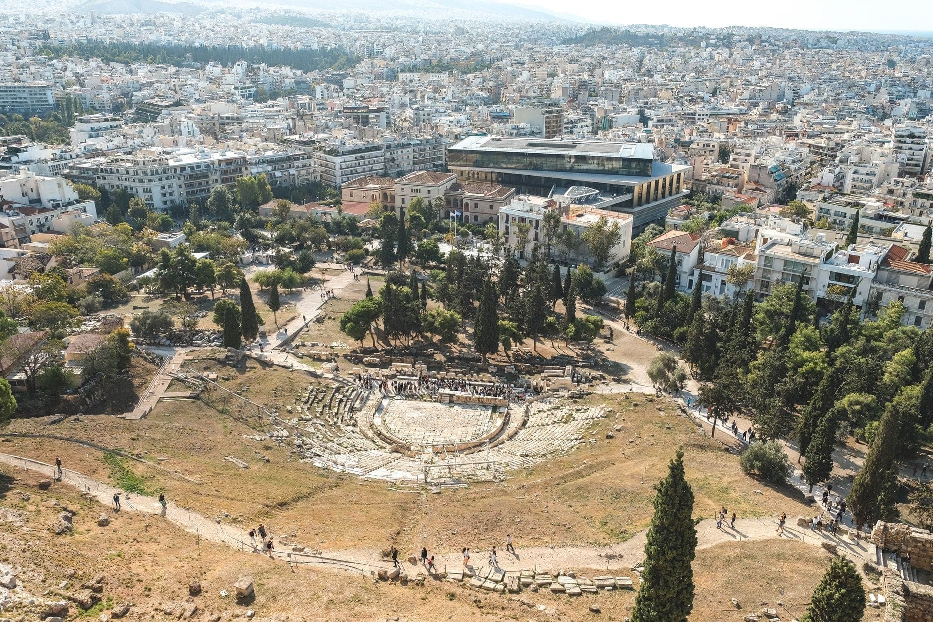 Aerial view of the ancient Theatre of Dionysus Eleuthereus on the Acropolis slope, surrounded by modern Athens and green trees under clear skies.