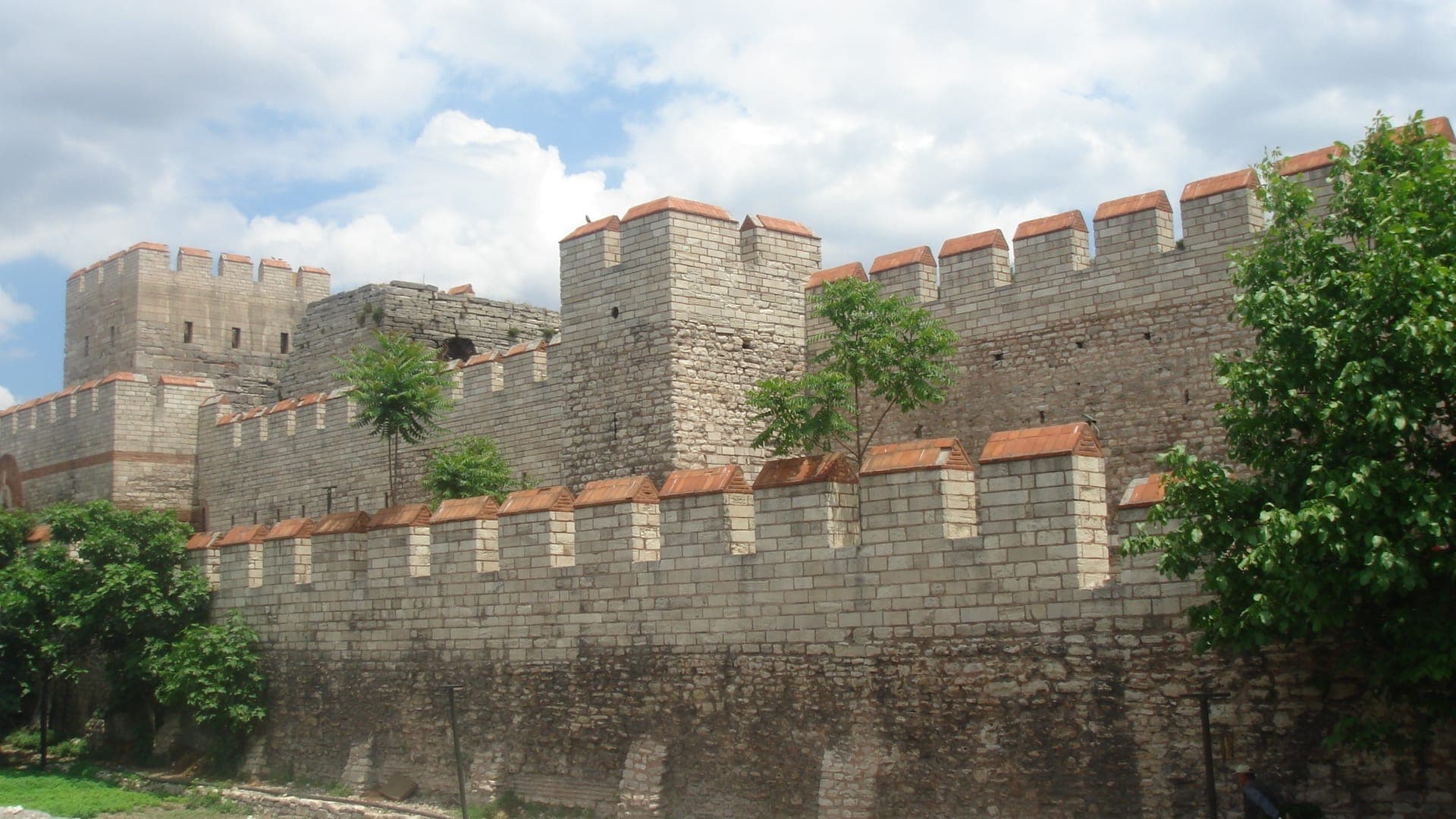 Steinwälle und Türme der Theodosianischen Mauern in Istanbul unter einem teilweise bewölkten Himmel, umgeben von Grünflächen.