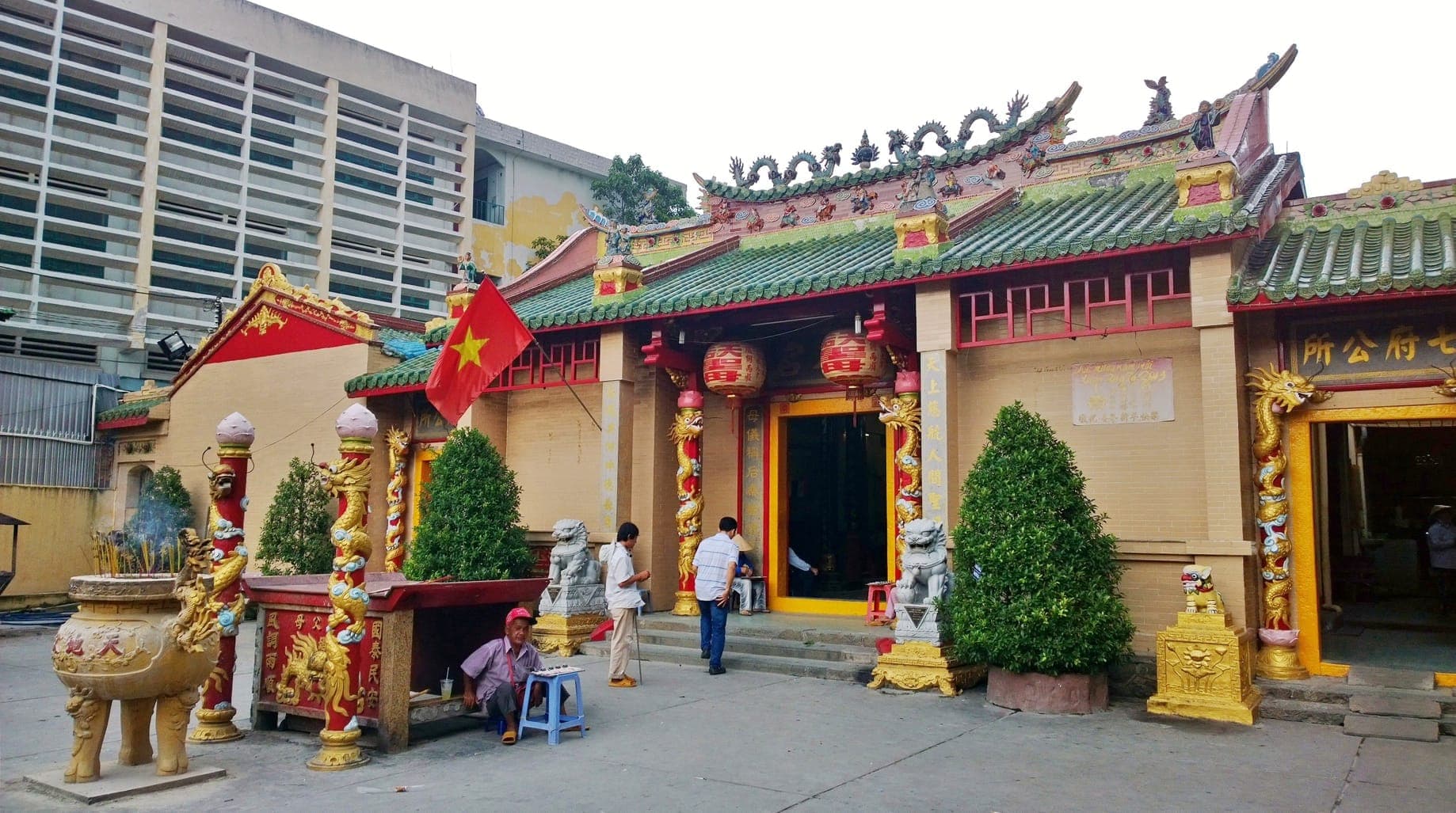 Lối vào chùa Thiên Hậu ở Chợ Lớn, nổi bật với các cột trụ rồng chạm khắc tinh xảo, tượng sư tử và du khách dưới bầu trời nhiều mây.