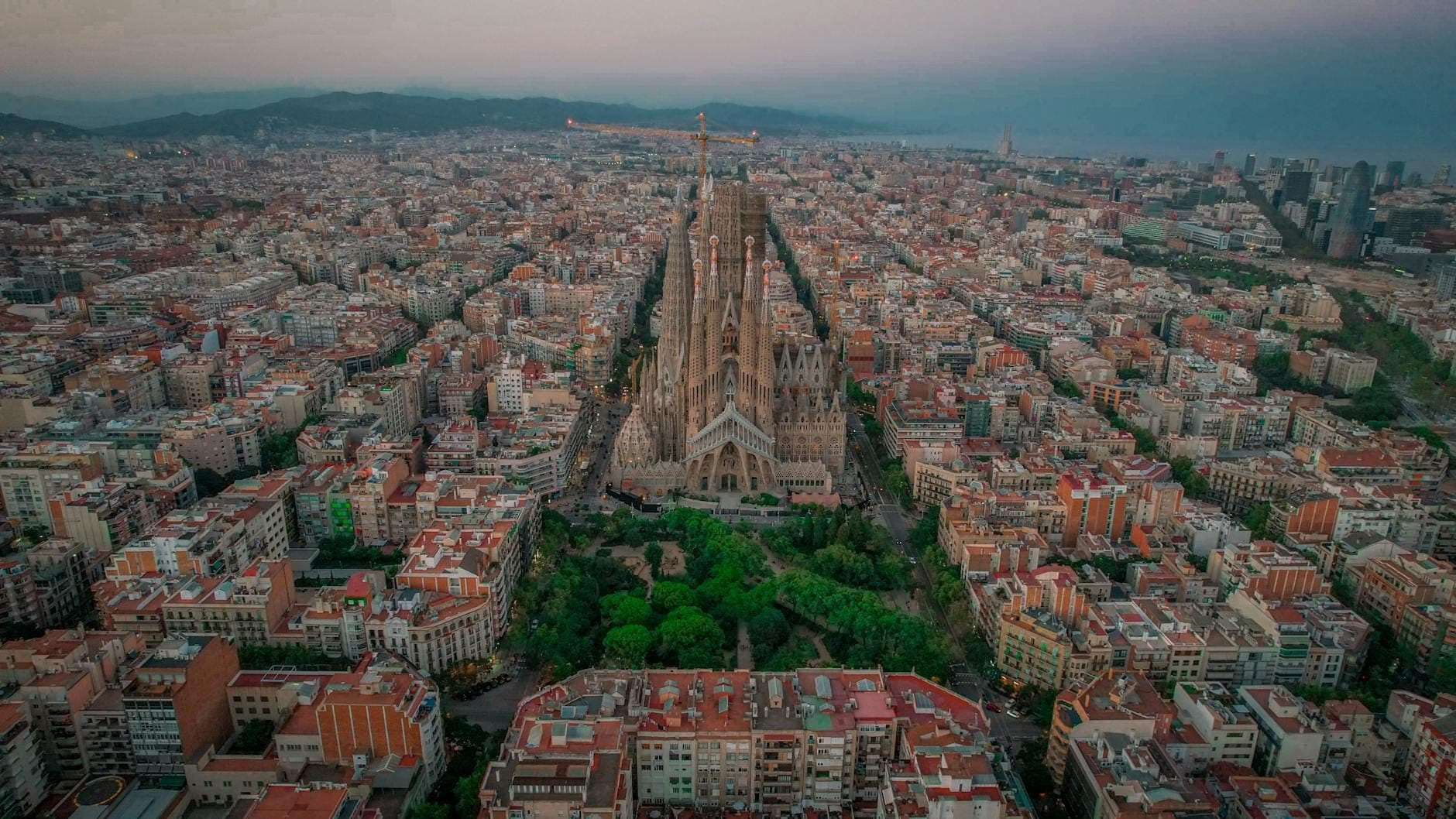 Toàn cảnh Barcelona nhìn từ trên cao, nổi bật với Sagrada Família uy nghi giữa những khu phố lúc hoàng hôn, xa xa là núi và biển.