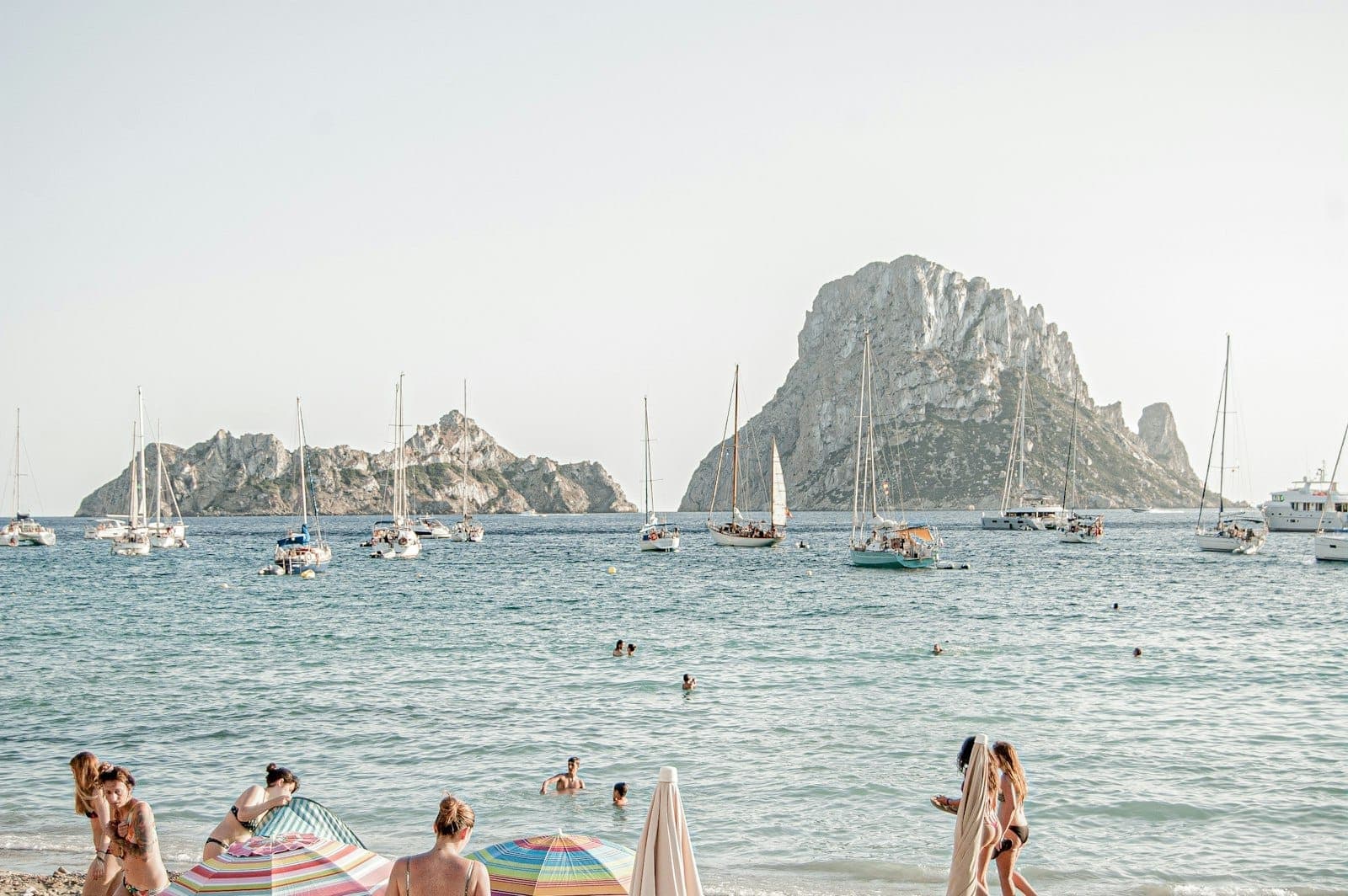 People relaxing under umbrellas on a sandy Ibiza beach, with sailboats anchored near Es Vedrà island and clear blue water.