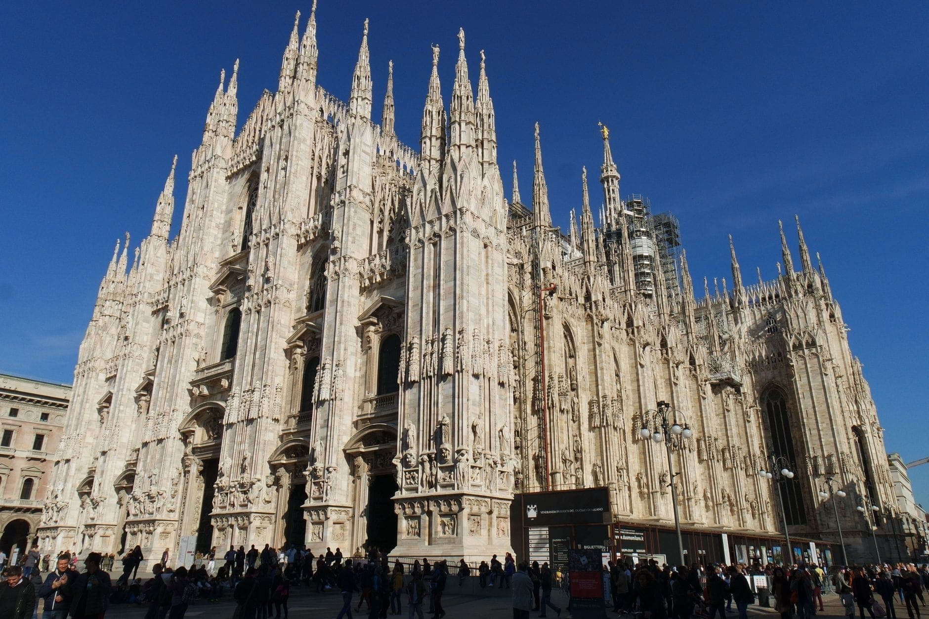 Vista amplia de la catedral Duomo de Milán con sus elaboradas agujas, fachada de mármol y multitudes en primer plano bajo un cielo azul despejado.