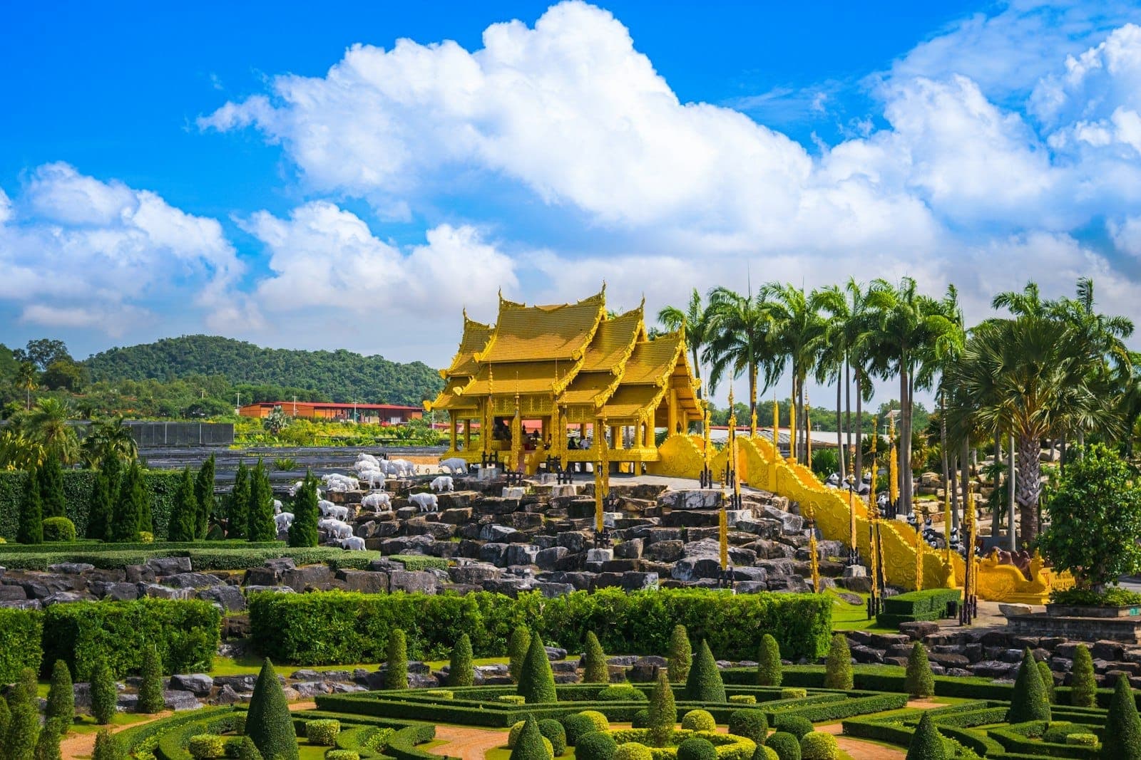 A beautifully landscaped garden with trimmed hedges, palm trees, and an ornate golden pavilion under a bright blue sky in Pattaya.