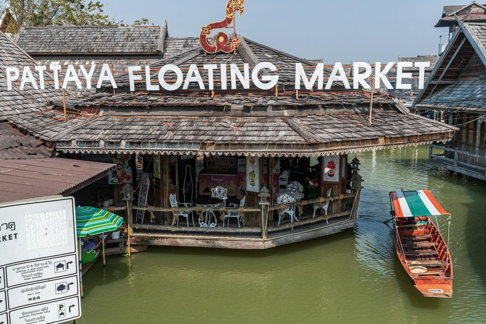 The Pattaya Floating Market features traditional wooden buildings above green water, with a colorful boat passing underneath its large white sign on a sunny day.