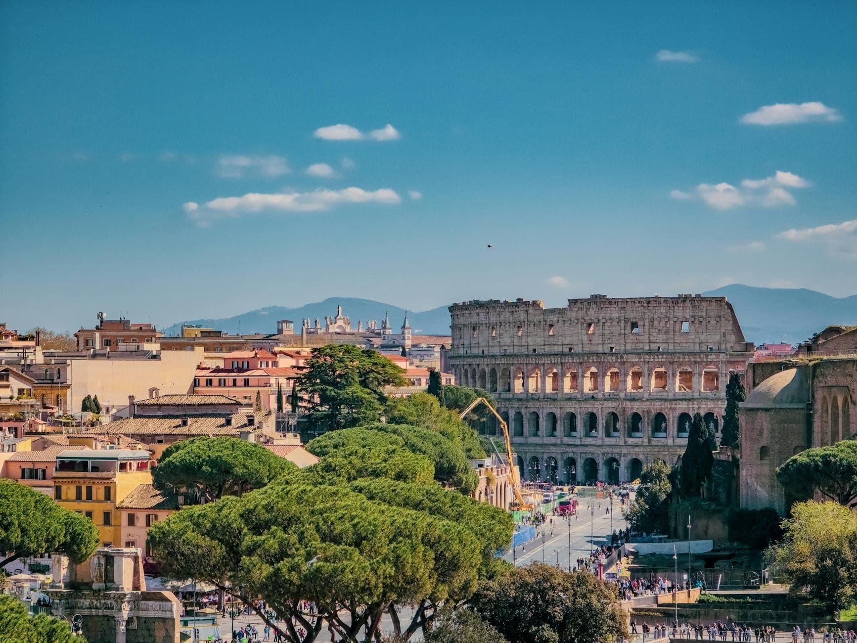 Toàn cảnh Rome với đấu trường Colosseum, những di tích cổ đại, mái nhà trải dài và dãy núi xa xa dưới bầu trời xanh trong, gợi lên không khí cổ điển đặc trưng của thành phố.