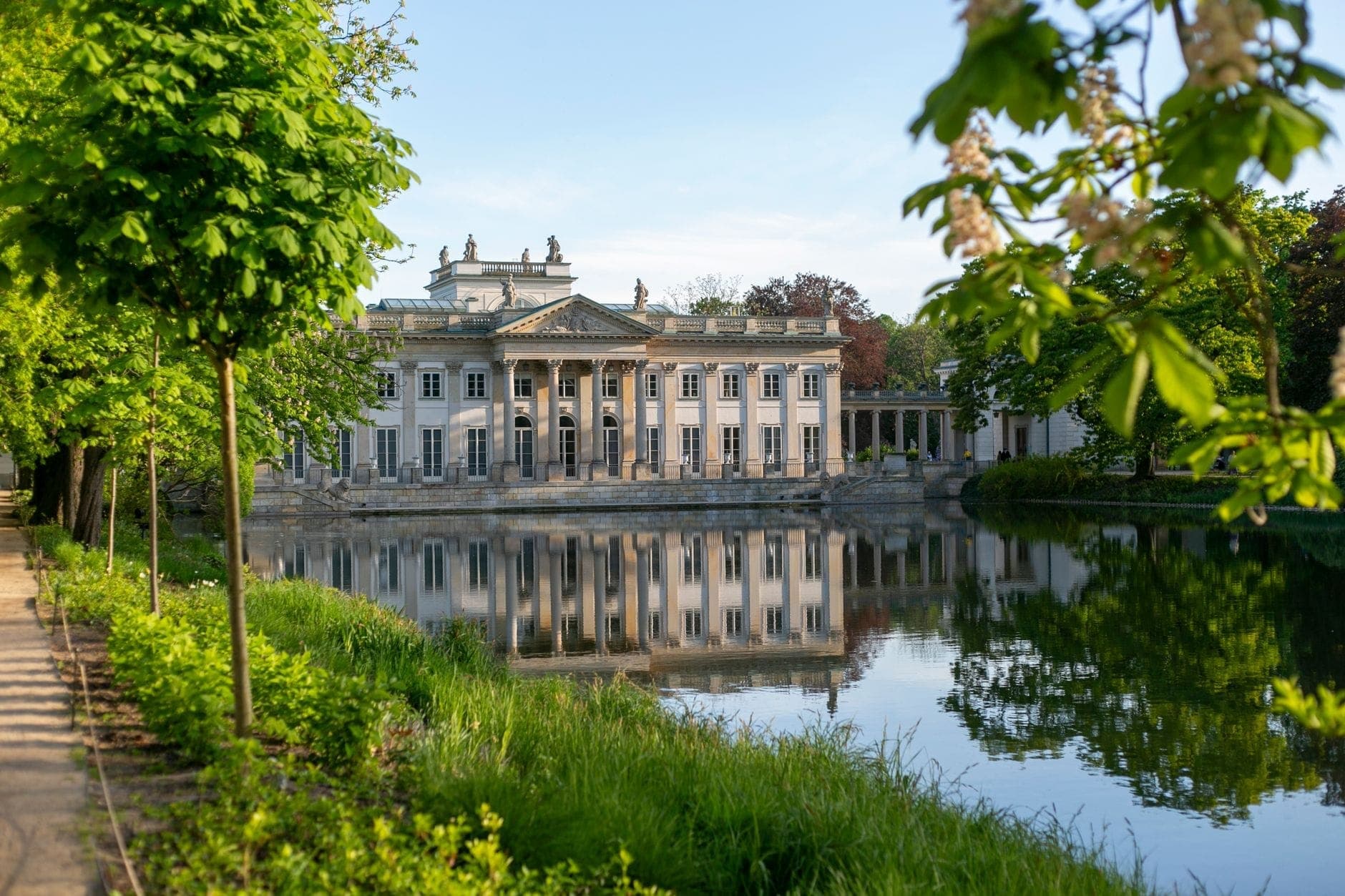 Tingin ng Łazienki Palace na nakasalamin sa tubig, napapalibutan ng malusog na mga puno at damo sa isang maliwanag na araw sa Warsaw.