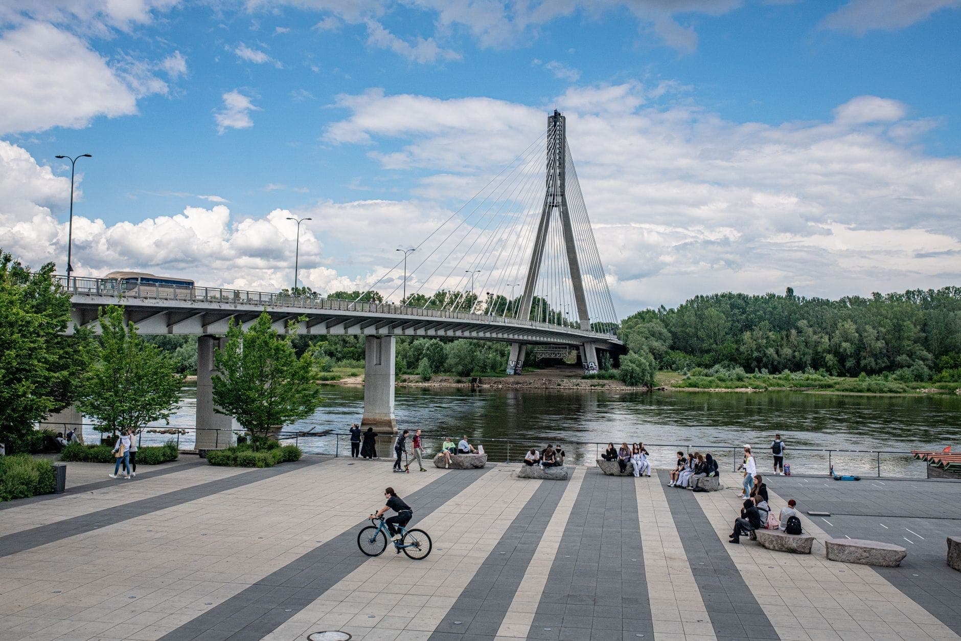 Promenade sa tabi ng ilog sa Warsaw na may mga taong nagpapahinga at nagbibisikleta malapit sa Tulay ng Świętokrzyski sa ibabaw ng Ilog Vistula sa isang maliwanag na araw.
