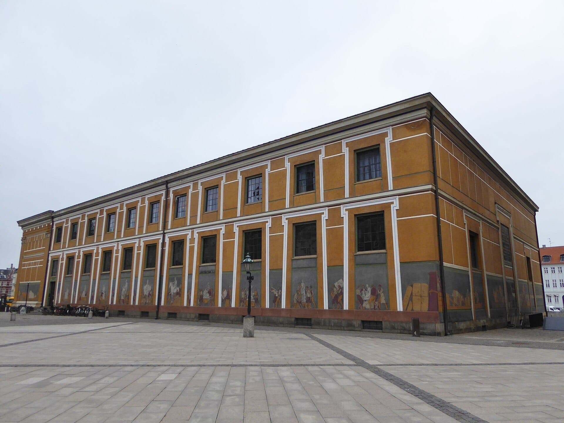 Außenansicht des Thorvaldsens Museum in Kopenhagen mit ockerfarbenen Wänden mit dekorativen Paneelen, hohen Fenstern und einem weitläufigen Kopfsteinpflasterplatz.