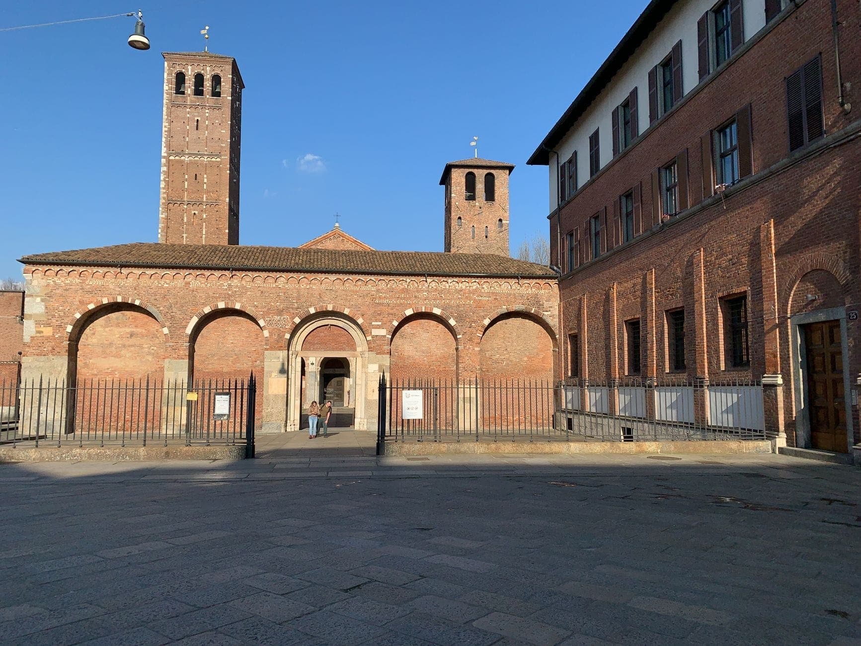 Basilica di Sant'Ambrogio in Milan with its Romanesque brick facade, arched portico, and twin bell towers under a clear blue sky.