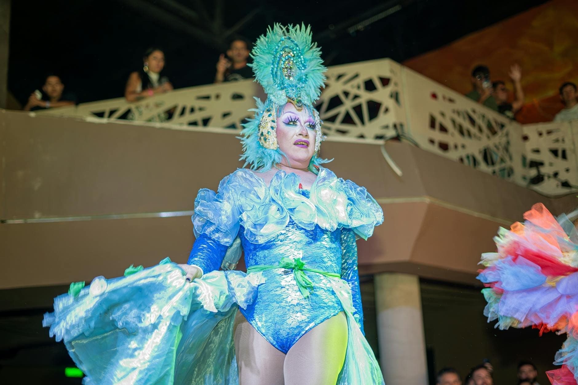 A vibrant cabaret performer in an elaborate blue feathered and sequined costume entertains audiences indoors at a lively show, with people watching from balconies in the background.