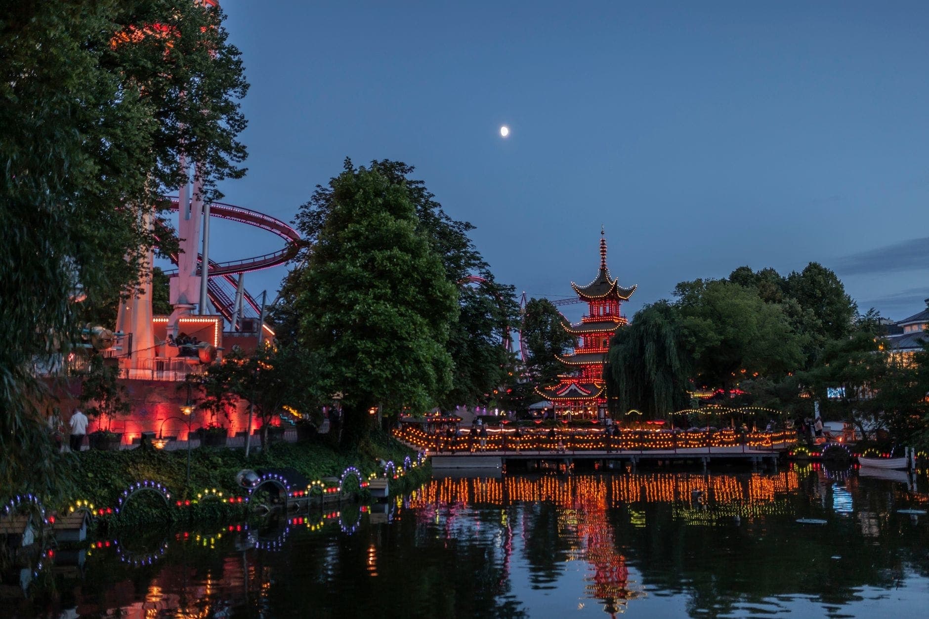 Abendlicher Blick auf den Tivoli Gardens mit einer beleuchteten Pagode am Seeufer, Laternen, Bäumen und Achterbahnschienen, die sich im Wasser unter einem Abendhimmel spiegeln.